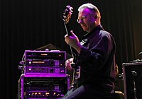 An older man from the side, holding a guitar with an amp to his left under a purple spotlight