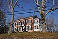 A brick house is framed by trees. A wooden two story addition on one side is painted white and has many windows. Otherwise the house trim is white with black shutters.