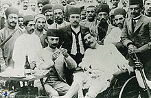 Doctor treating an injured man, with a crowd of standing men behind them