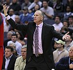 A man, wearing a black suit and a tie, is making a gesture by raising his hands while standing on the side of a basketball court.