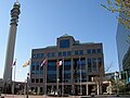 Une photographie de l'hôtel de ville de Moncton, montrant une rangée de cinq drapeaux sur son parvis. De la gauche à la droite se trouvent le drapeau du Canada, le drapeau du Nouveau-Brunswick, le drapeau du Royaume-Uni, le drapeau de l'Acadie et le drapeau de Moncton.