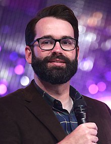 close-up of Matt Walsh wearing a dark checkered shirt and dark blazer, holding a microphone, looking right of camera, with blurry lights in the background