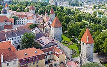 Farbfotografie in der Obersicht von Häusern und mittelalterlichen Wachtürmen mit braunen Dächern, die quer von unten rechts nach oben links gehen. Zwischen den beiden vorderen Türmen ist eine kleine Steinmauer. Im Hintergrund sind eine Grünanlage und moderne Gebäude.