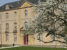 Façade d'un bâtiment austère à deux étages et un étage mansardé. Un arbre en fleurs au premier plan.