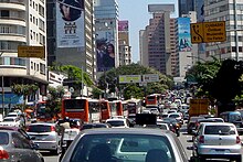 Looking down a busy road, which is banked on both sides by tall buildings, some of which are covered in advertisement billboards