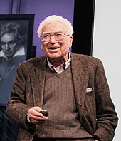 Half-length portrait of a white-haired man in his seventies talking. A painting of Beethoven is in the background.