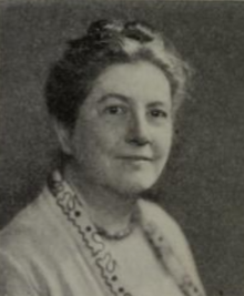 A white woman with hair in an updo, wearing glasses and a soft light-colored jacket