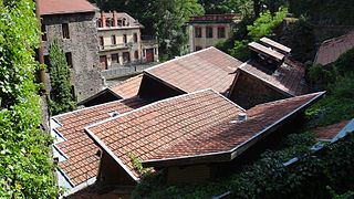 Vue sur un ensemble de toits en tuiles mécaniques constitutifs des bâtiments de la forge Mondière puis au fond 2 grandes bâtisses industrielles