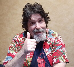 Headshot van Mick Foley met zwart haar en een grijze baard. Foley kijkt recht in de camera, steekt zijn rechterduim omhoog en draagt een zwart T-shirt met daaroverheen een vrolijk gekleurd rood overhemd.