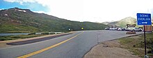 A road going through a mountainous area with no trees. On the left is a small pond. On the right is a parking lot with two buildings. A brown and yellow sign in the distance in the parking lot reads "Independence Pass ... Contintental Divide" while a blue one on the very right says "Scenic Overlook" in white with an arrow pointing to the right