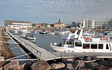 Plusieurs bateaux de pêche sont amarrés le long d'un quai. À l'arrière plan, trois édifices se dressent au loin.