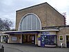 A beige-bricked building with a dark blue, rectangular sign reading "LOUGHTON STATION" in white letters all under a blue sky with white clouds