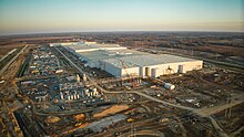 Photo is of a large scale manufacturing facility under construction, captured from above with aerial photography. There are many cranes, heavy equipment, materials, and other machinery surrounding the tall plain walls of the main facility.