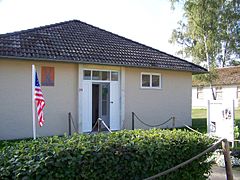 Un petit pavillon de plain-pied. Devant l'entrée, une haie et un drapeau américain. Un panneau annonce qu'il s'agit de la maison de Jesse Owens ; le reste du texte est illisible en raison de la luminosité trop haute.