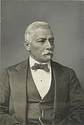 Portrait d'un homme aux cheveux gris, en costume et nœud papillon.