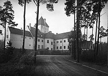 A large white building with a dark coloured roof. The building is surrounded by a number of trees.