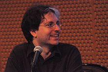 A photograph of Douglas Wolk, a white man with unkempt hair and a warm smile, sitting behind a conference table.