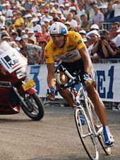 A man on a bicycle wearing a helmet with a visor and a yellow top with black shorts. In the background, there is a red motorcycle with two men on it.