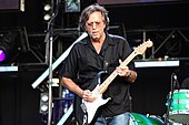 A man on a stage wearing blue jeans, a black shirt and eyeglasses. He is holding a black guitar, and part of a drum set and other audio and stage equipment can be seen in the background.