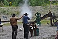 A Cowboy action shooter firing a lever action rifle at steel targets. The Range Officer to the left is holding a shooting timer to measure the time.