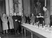 Discours d'un homme politique debout qui lit une feuille écrite, lors d'un banquet ; un groupe d'hommes l'écoutent.