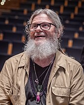 A bearded man with glasses wearing a brown jacket over a black shirt and several chains.