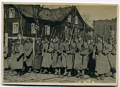 Photographie en noir et blancs de soldats de l'Armée rouge défilant alignés, tenant chacun un fusil, avec derrière eux un bâtiment en bois.