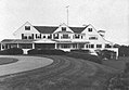 A large white house with three large gables and a porch running for much of its width stands before an expansive lawn and curved drive.