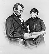 A seated Lincoln holding a book as his young son looks at it