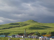 Photographie d'une colline entièrement verte, avec en bas des maisons au premier plan.