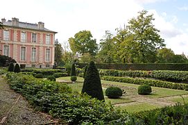 Le jardin clos Art déco vu légèrement de côté avec la Folie en arrière-plan à gauche. À gauche (1er plan) une allée décorée de briques, au centre une pelouse entouré de buissons taillés.