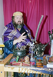 A middle-aged white man with a grey beard sits, staring at the viewer. In front of him is a table with various objects placed upon it, including a small cauldron with large iron nails protruding from it.