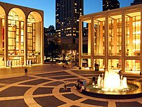 The corner of a lit up plaza with a fountain in the center and the ends of two brightly lit buildings with tall arches on the square.