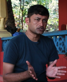 Half-length portrait of Kureshi speaking in front of a red building and some trees