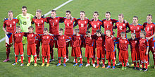 Photographie de face de onze joueurs de football au maillot rouge, derrière onze enfants.