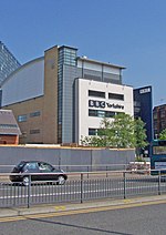 A quirky modern five-storey building with a large sign saying "BBC Yorkshire" in black above the second-floor windows on the white-fronted facade of the lower four floors can be seen on the far side of a dual-carriageway road with a barrier along the central reservation. At right-angles to the right of the building is a tall blue slab with the letters "BBC" in white at the top. The left side of the building is mostly brick-red with a few windows, but above it is a light blue windowless section. The roof above this and the grey fifth floor of the frontage curves gently down to the rear. A lone car is driving from left to right along the road; between it and the building, temporary boards have been erected in front of a building to the left. In the top left-hand corner of the picture, part of a tall many-windowed building can be seen.