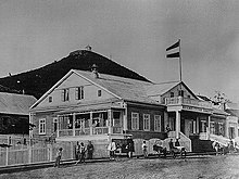 Photographie en noir et blanc d'une maison avec un étage qui fait flotter le pavillon de l'Empire russe. La maison, en bois, est sur le bord d'une rue assez large, et au fond se distingue une montagne.