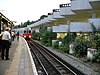 A group of people standing on a railway platform with a railway track to the right and a white-and-red train running on it with its headlights on