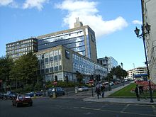 A road-junction with traffic-lights, traffic queuing uphill and lamp-standards with pairs of lights. In the foreground on the right is part of a flower-bed, and behind it, uphill in the distance, is a multi-storey car-park. On the left is a complex of four twentieth-century buildings flanked by a number of roadside trees: at the front is a three-storey building with bands of windows and some light brown stone cladding, while behind it to the right is a much taller end-on block in the same style; to the left of this block, an older building of the same height with alternate bands of concrete and windows; further up the road, a more recent lower-rise sleek white building.