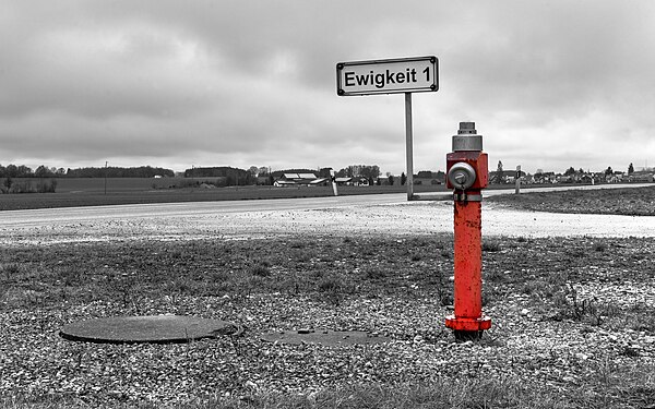 Red hydrant, standing in drizzling rain under a brooding sky in Lengenfeld, Germany. The sign reads "Eternity 1".