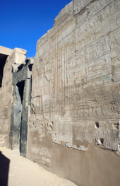 Ground-level outside view of stone walls with raised-relief carvings of human figures and hieroglyphic writing; a doorway is positioned at the center; the top left portion shows a blue sky without clouds.