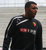 A young man wearing a black and white top. The logo on his top is coloured red, black and yellow, and reads "WATFORD"