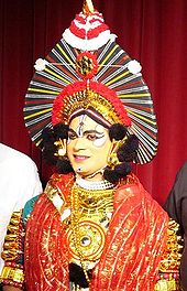 Person with painted eyes in Yakshagana costume, as gold-spangled robe with red sheer scarf and spiked headress on gold crown