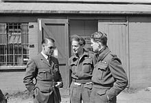 Photographie noir et blanc de trois aviateurs en uniforme, les mains dans les poches, discutant.