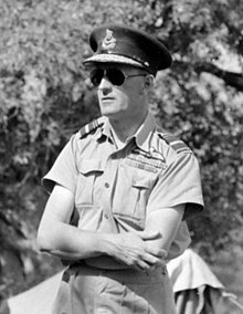 Portrait en noir et blanc d'un homme vêtu d'un costume militaire se tenant près d'un arbre.