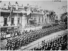 Photographie d'un défilé militaire avec des troupes, passant devant un bâtiment pavoisé des drapeaux (de gauche à droite) américain, français, anglais, et de l'Empire japonais.