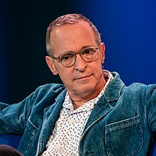 A bust photograph of a white man in spectacles; he is wearing a white patterned shirt, blue jacket, and a jaw-mounted microphone. He is facing the camera, looking to its left.