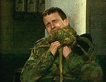 A man in camouflage fatigues winces with pain as he tries to remove a green alien creature from his neck.