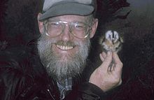 A smiling, bearded man holds a small bird near his left ear with his right hand.