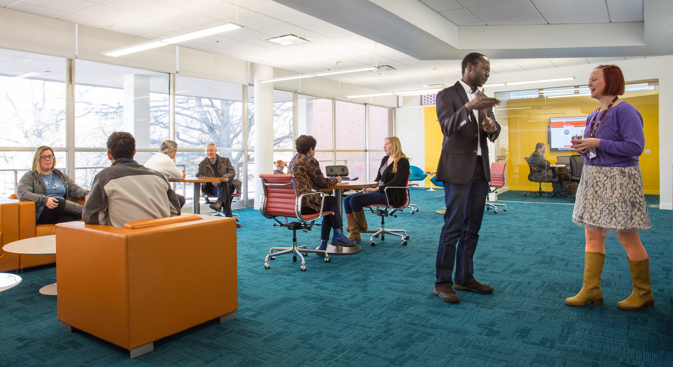 People chatting in a colorful room with large windows, a big screen, and cool chairs